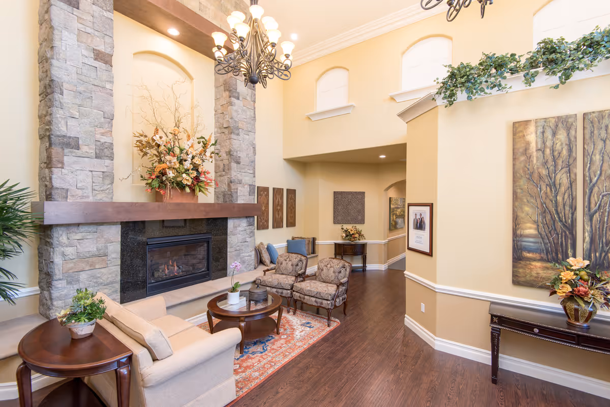 A cozy living room area with a stone fireplace, a beige sofa, two patterned armchairs, a round wooden coffee table, and decorative plants and flowers. The room has high ceilings with a chandelier, light yellow walls, and hardwood floors. Artwork and framed pictures adorn the walls.