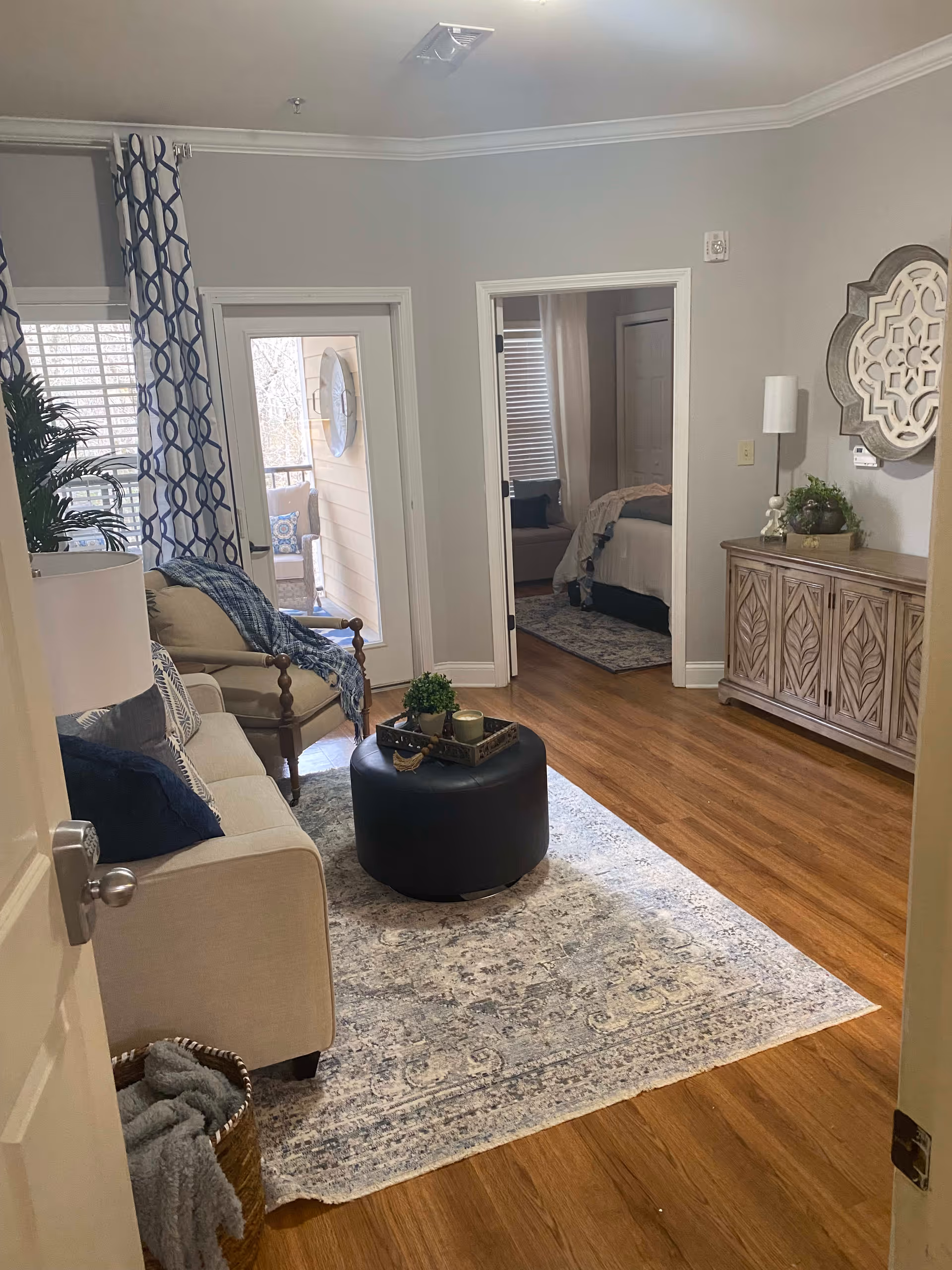 A cozy living room with a beige sofa and armchair, a round black ottoman with a tray holding candles and a small plant, a patterned rug on wooden flooring, and a wooden sideboard with decorative items and a wall mirror. There is a doorway leading to a bedroom and a glass door opening to a balcony with outdoor seating.