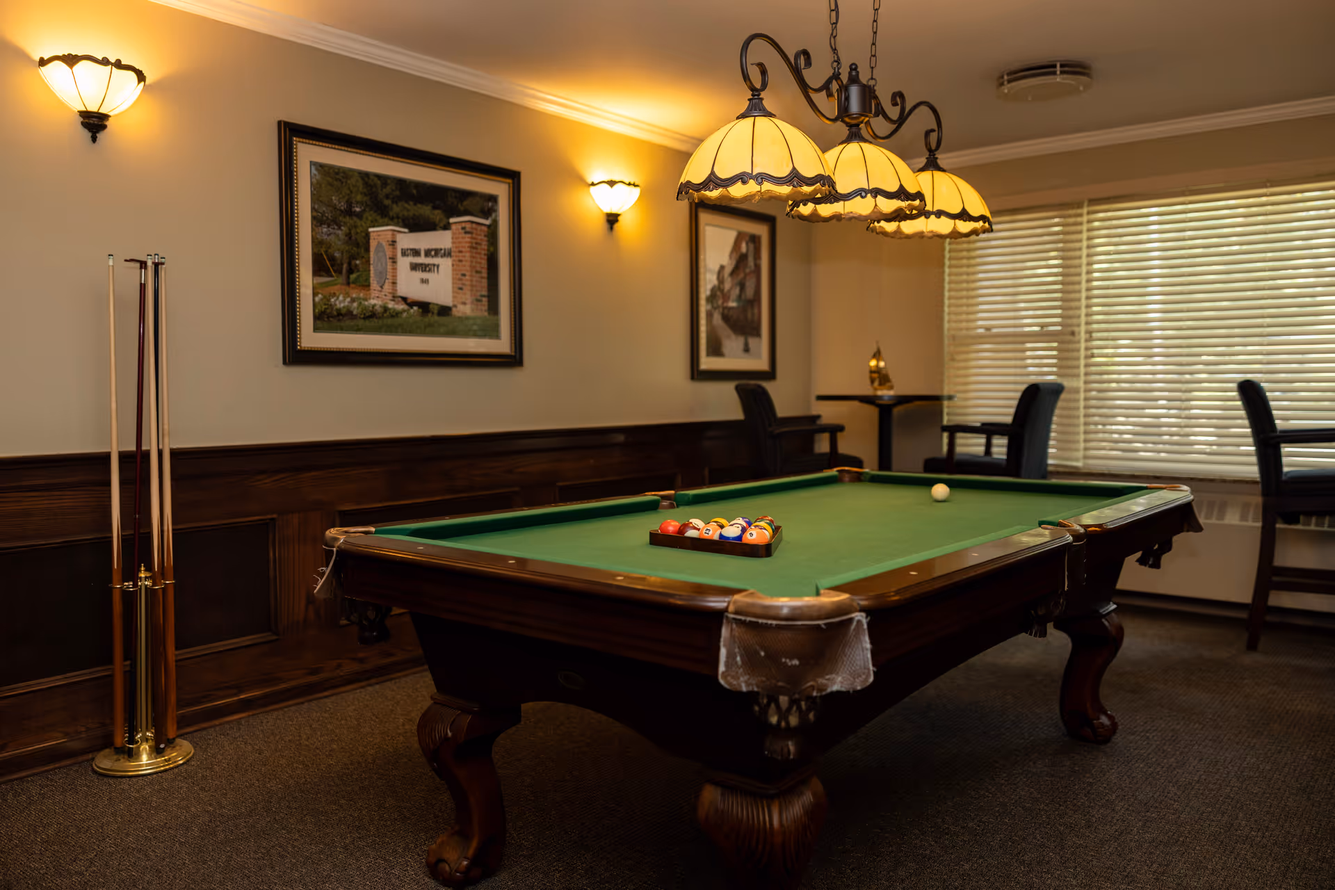 A cozy room with a green felt pool table in the center, pool cues standing in a rack to the left, and three hanging lamps above the table. The room has wood paneling on the lower half of the walls, framed pictures on the walls, and a window with blinds on the right side. There are also a few chairs and a small table near the window.