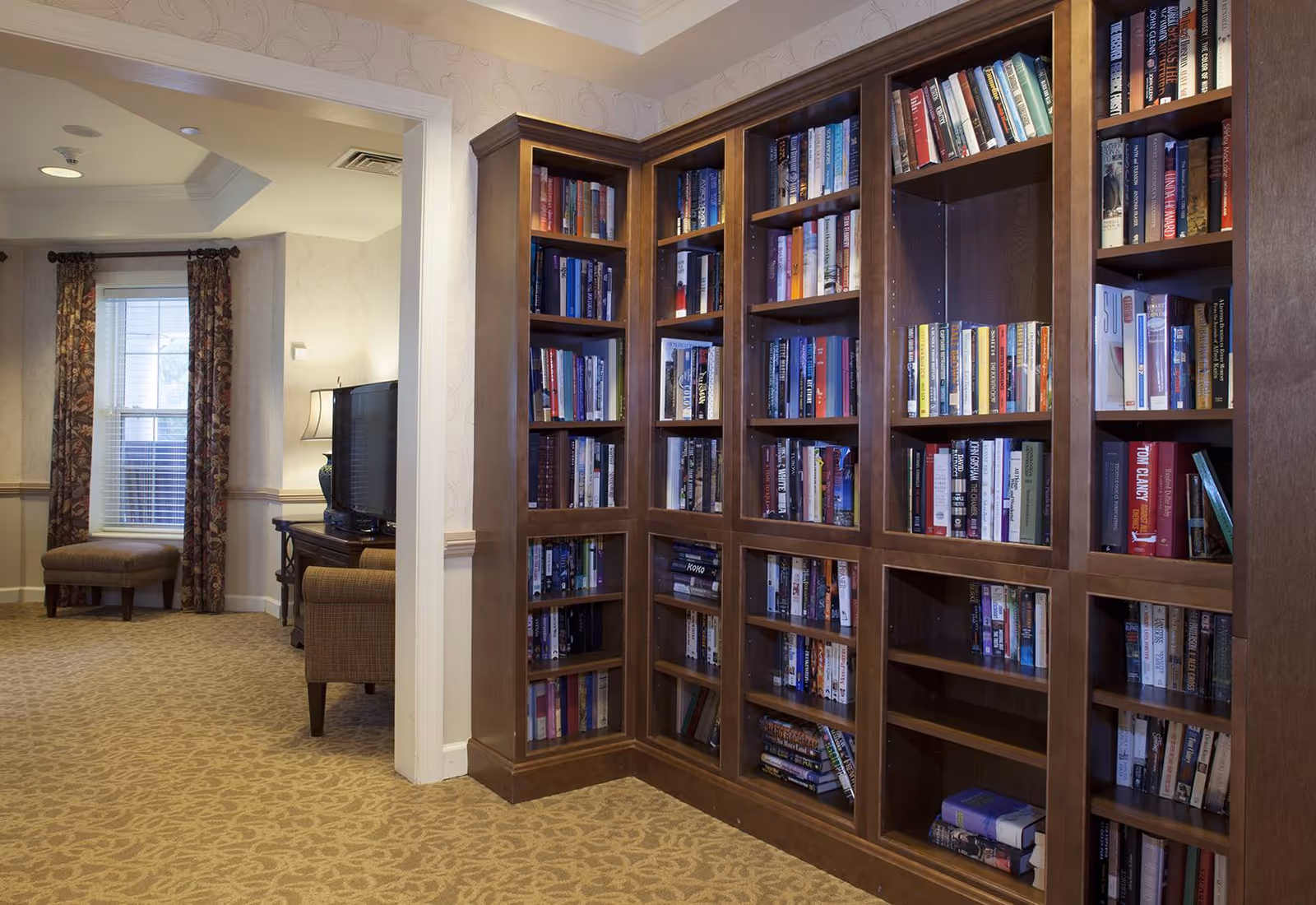 Built-in wooden bookshelves filled with books in a cozy common room with seating and a TV visible through a doorway.