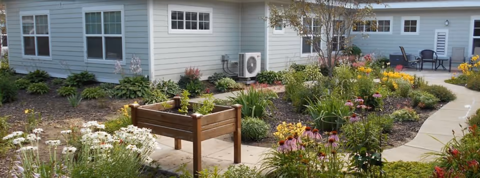 A landscaped garden area with various flowering plants and shrubs surrounding a paved walkway. There is a raised wooden planter box in the foreground and patio furniture with chairs and a table in the background near a light blue building with multiple windows.