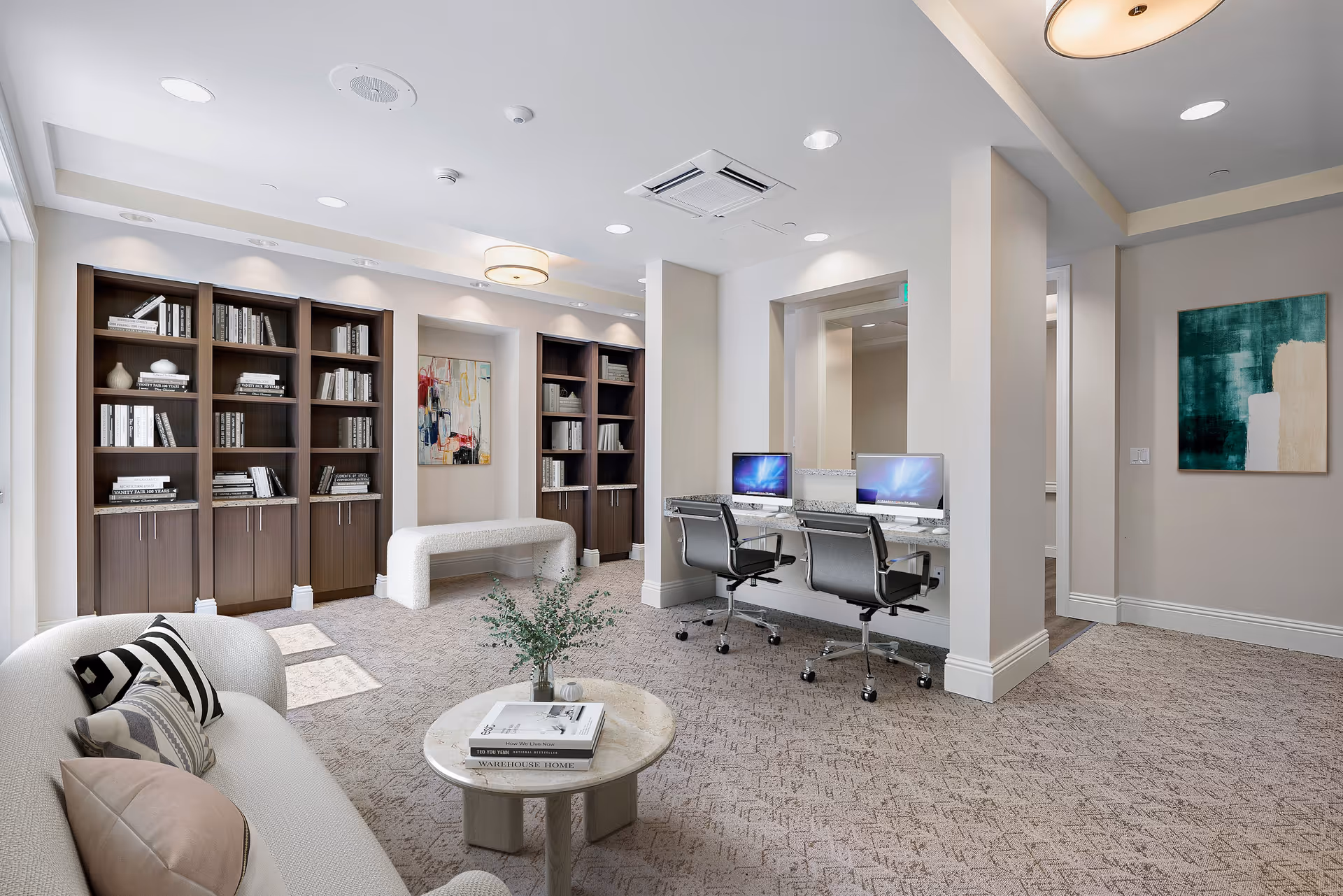 A modern and bright interior room featuring a cozy seating area with a white sofa and patterned pillows, a round marble coffee table with books and a small plant, built-in dark wood bookshelves filled with books and decorative items, two computer workstations with office chairs, and abstract artwork on the walls. The room has carpeted flooring and recessed lighting.