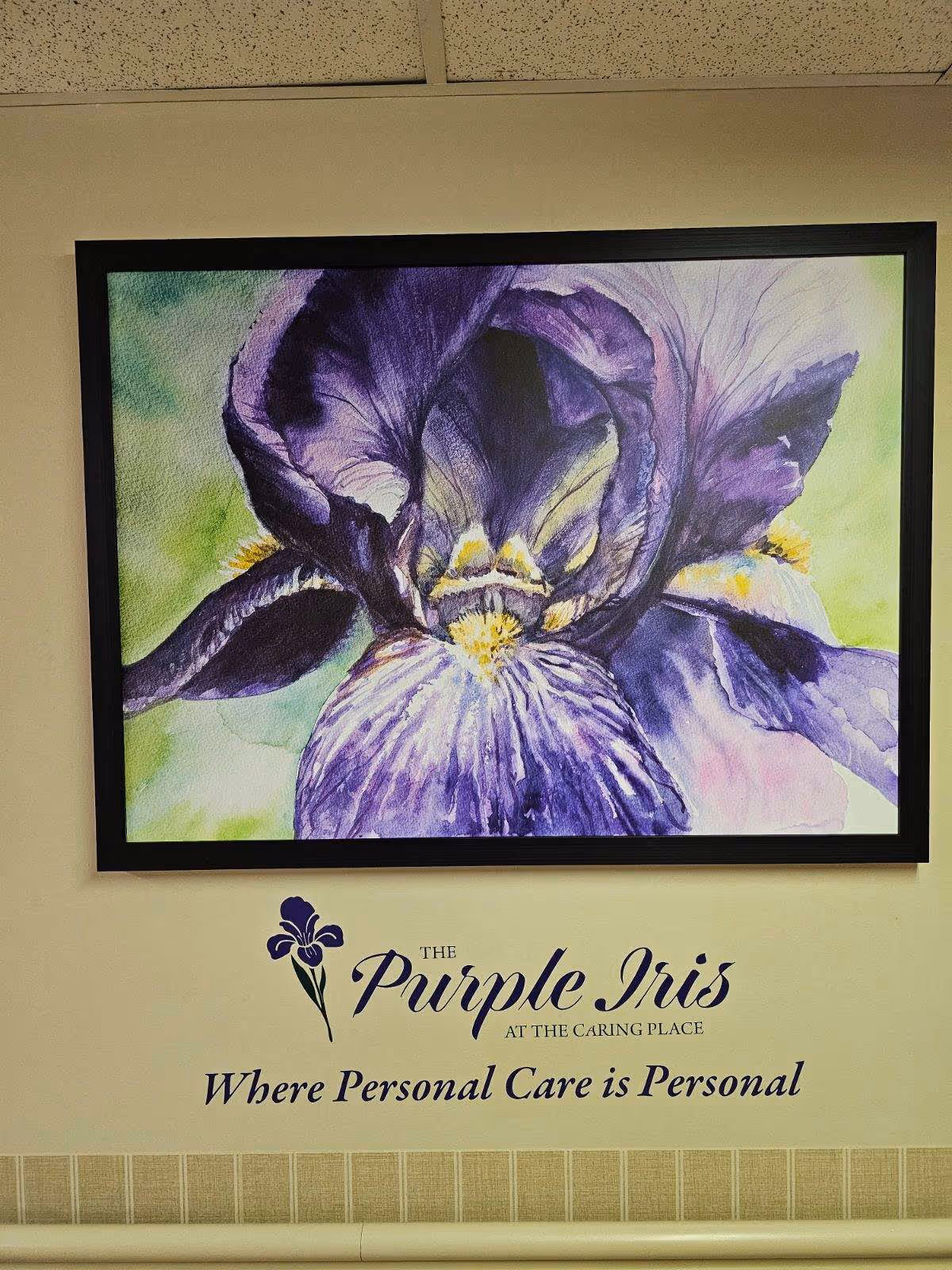 Framed purple iris painting hung above wall lettering that reads 'The Purple Iris at The Caring Place — Where Personal Care is Personal'.