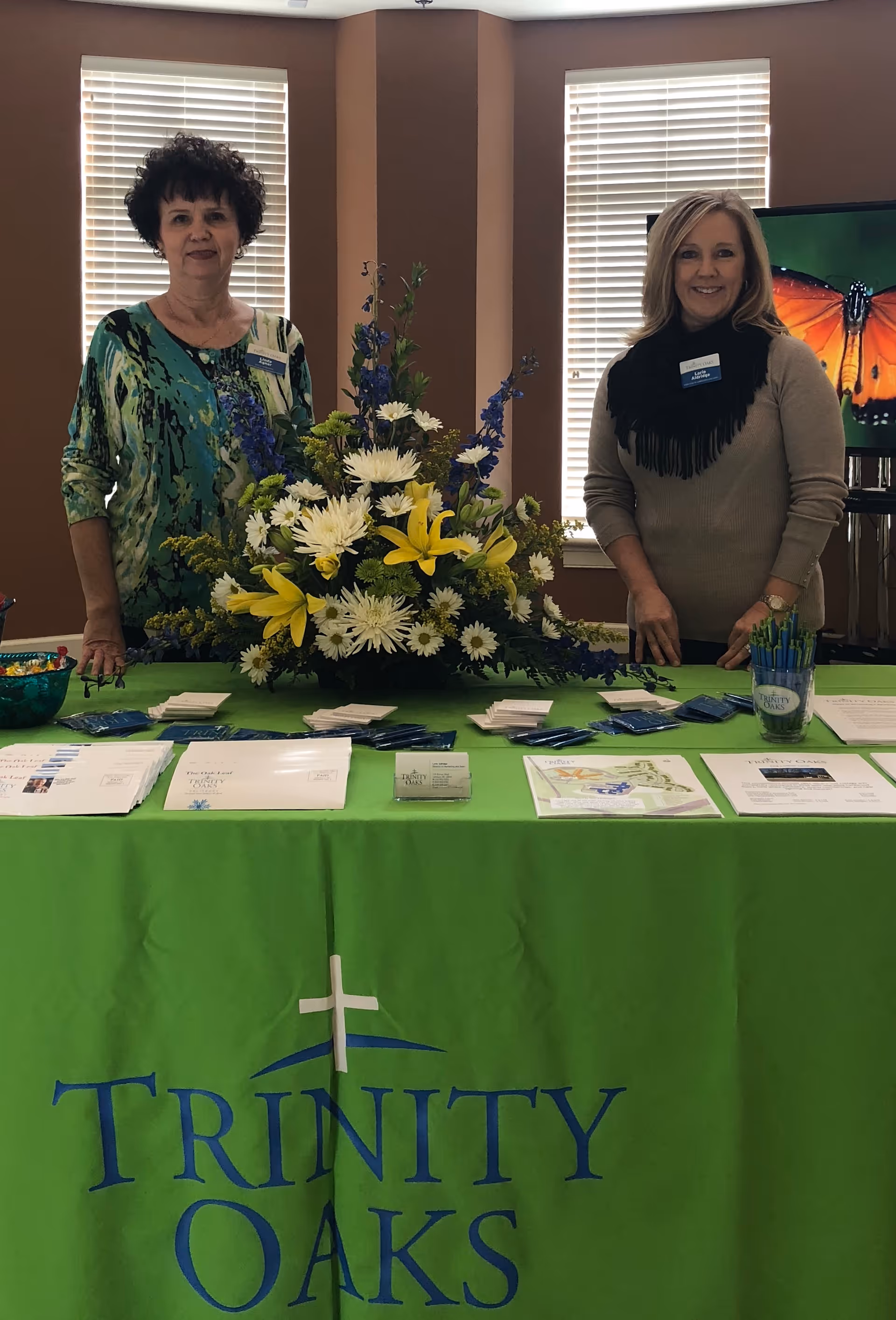Two women standing behind a table covered with a green tablecloth that has the Trinity Oaks logo. The table displays brochures, business cards, pens, and a large floral arrangement with yellow and white flowers. The background shows two windows with blinds and a TV screen with a butterfly image.