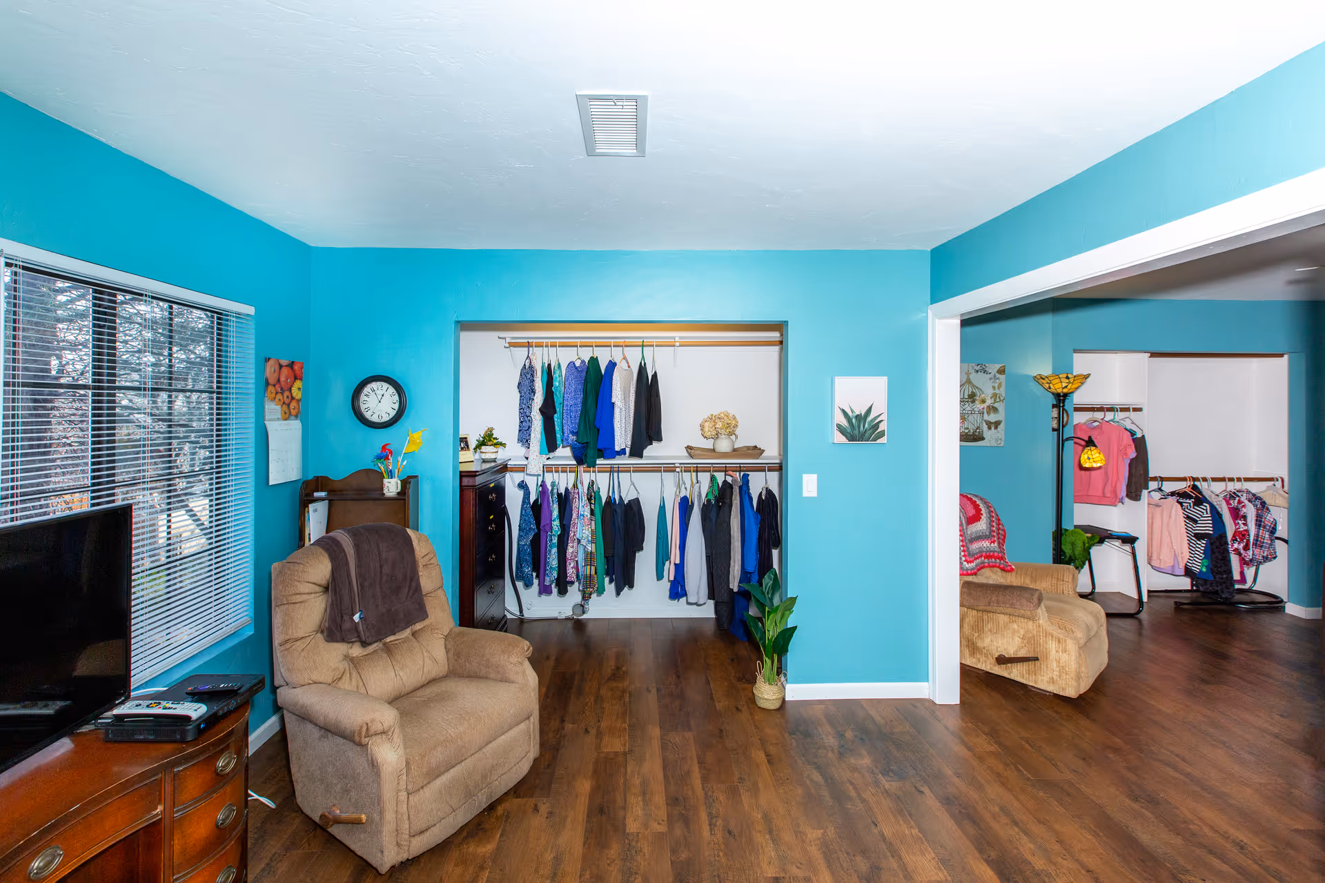 Interior view of a living space with turquoise walls and wooden flooring. There are two beige recliner chairs, one near a window with blinds and a TV on a wooden dresser, and another chair in an adjacent room. Both rooms have open closets with clothes hanging. The space is decorated with a wall clock, a small plant, and framed pictures.