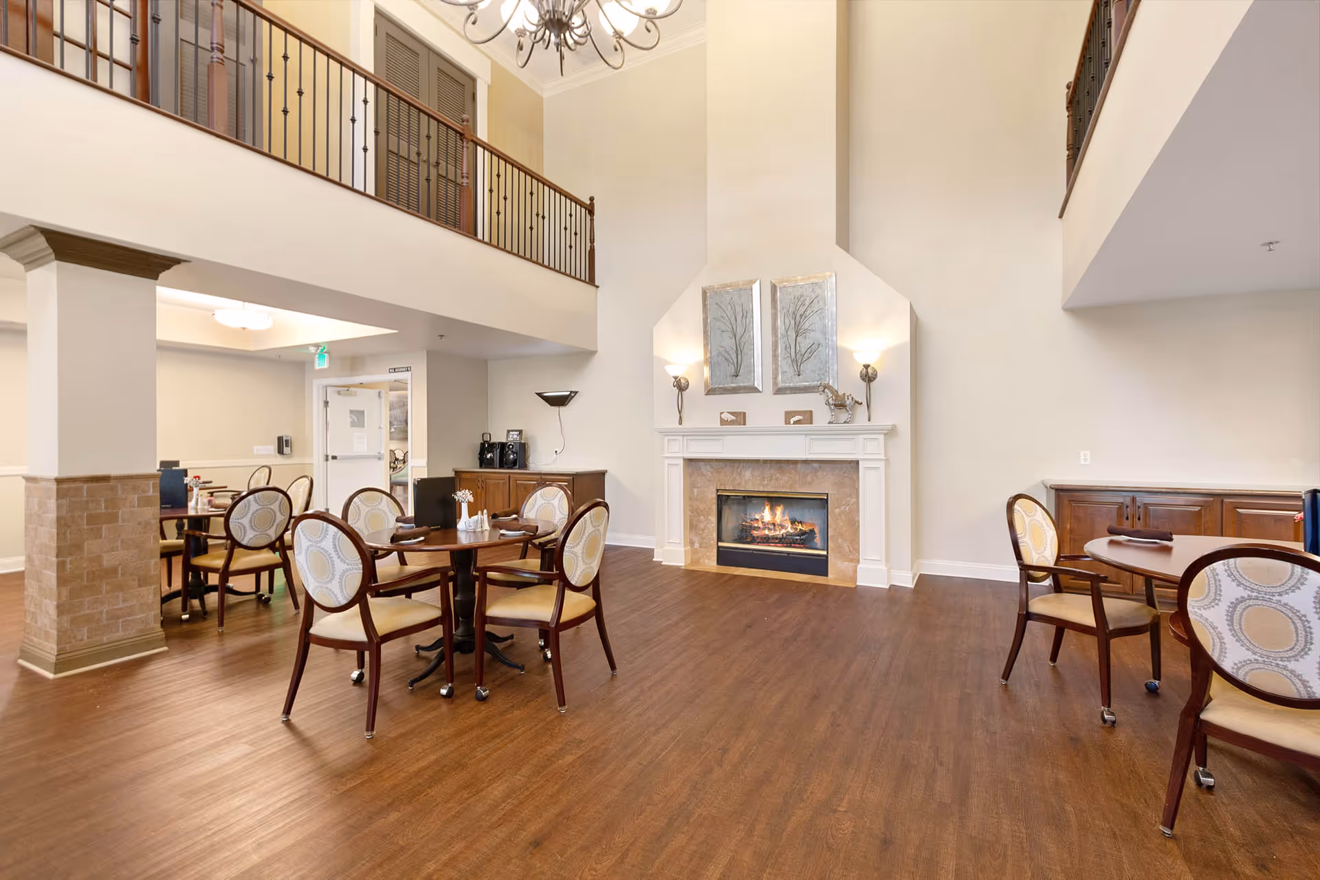 A spacious and well-lit common area with wooden flooring, several round tables with cushioned chairs, a lit fireplace with decorative artwork above it, and a balcony railing on the upper level.