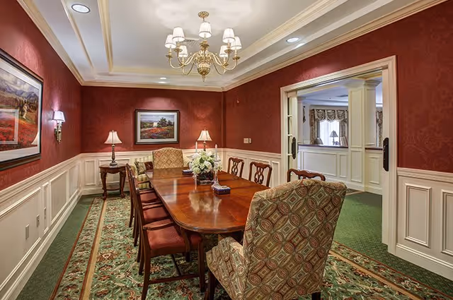 A formal dining room with a long wooden table surrounded by upholstered chairs. The walls are decorated with red wallpaper and white wainscoting, and there are framed landscape paintings on the walls. The room is lit by a chandelier and table lamps, and there is a patterned green carpet on the floor. An adjacent room with white paneling and windows is visible through a wide doorway.