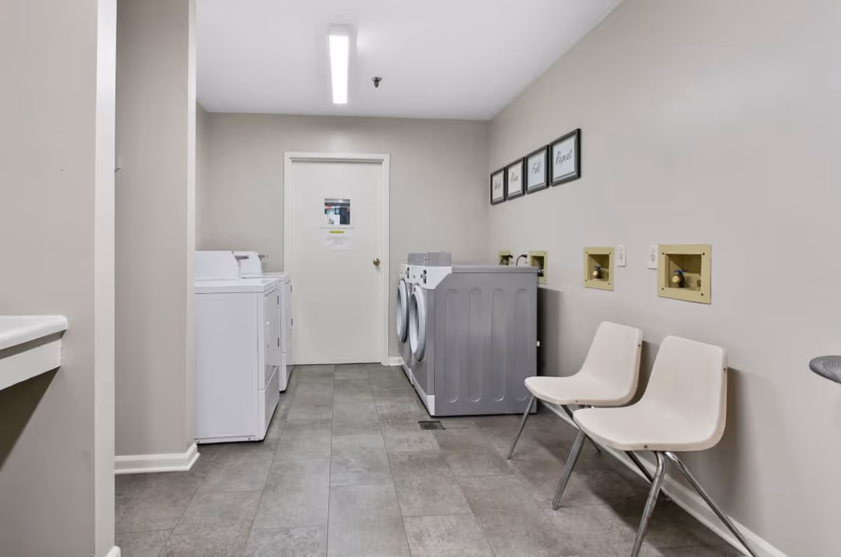 Laundry room with two white washing machines and two white dryers along the left and back walls. Two white plastic chairs with metal legs are placed against the right wall. The walls are painted light beige, and there are four framed pictures with words on the right wall above the chairs. The floor is covered with gray tiles, and a white door is visible at the back of the room.