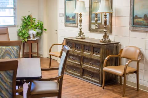 Communal dining area with a wooden table and chairs, a decorative sideboard topped with lamps and framed artwork, and a potted plant near the window.