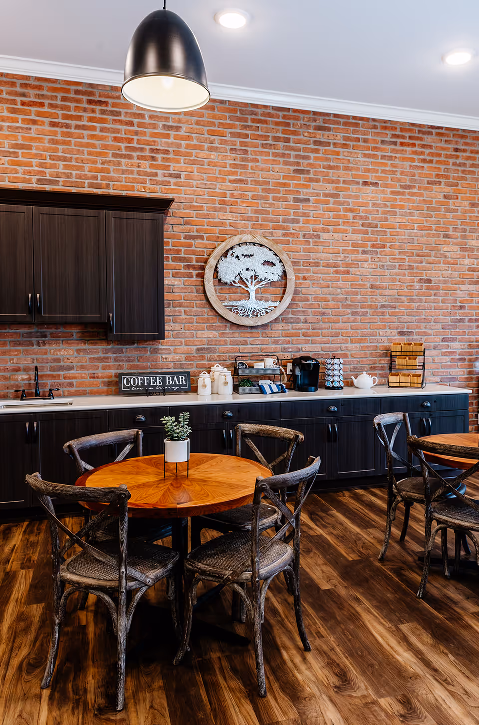 Cozy communal dining area with round wooden tables and chairs, a coffee bar along a brick wall, and pendant lighting.