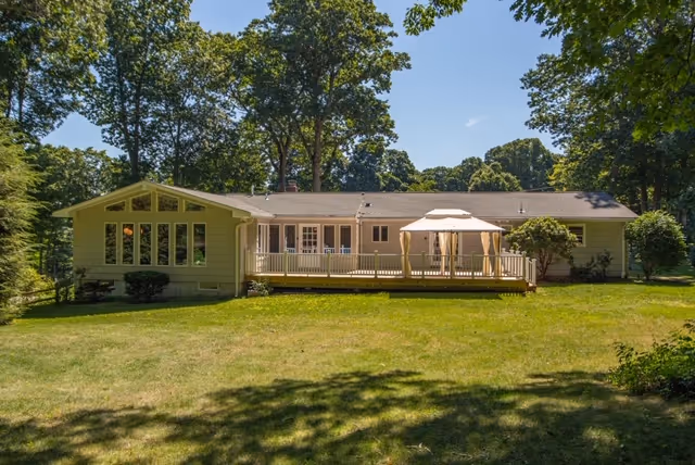 Single-story residential building with a large wooden deck featuring a canopy-covered seating area, surrounded by a grassy yard and tall trees under a clear blue sky.