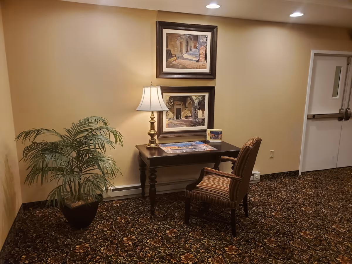 A small seating area with a wooden desk and a striped upholstered chair on a patterned carpet. On the desk is a brass table lamp, a puzzle, and a small framed picture. Two framed paintings hang on the beige wall above the desk. A potted plant is placed to the left of the desk, and a white double door is visible on the right side of the image.