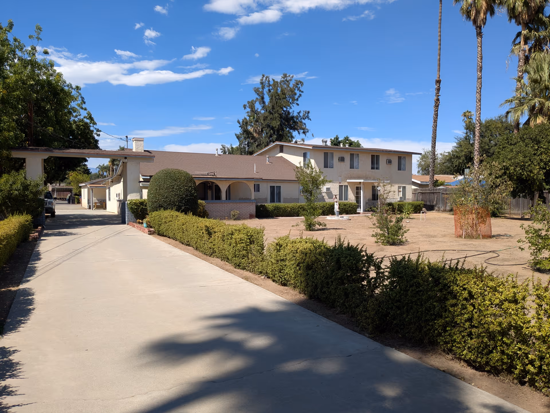 Driveway leading to a two-story beige residential building with trimmed hedges, a dry front yard and tall palm trees under a blue sky.