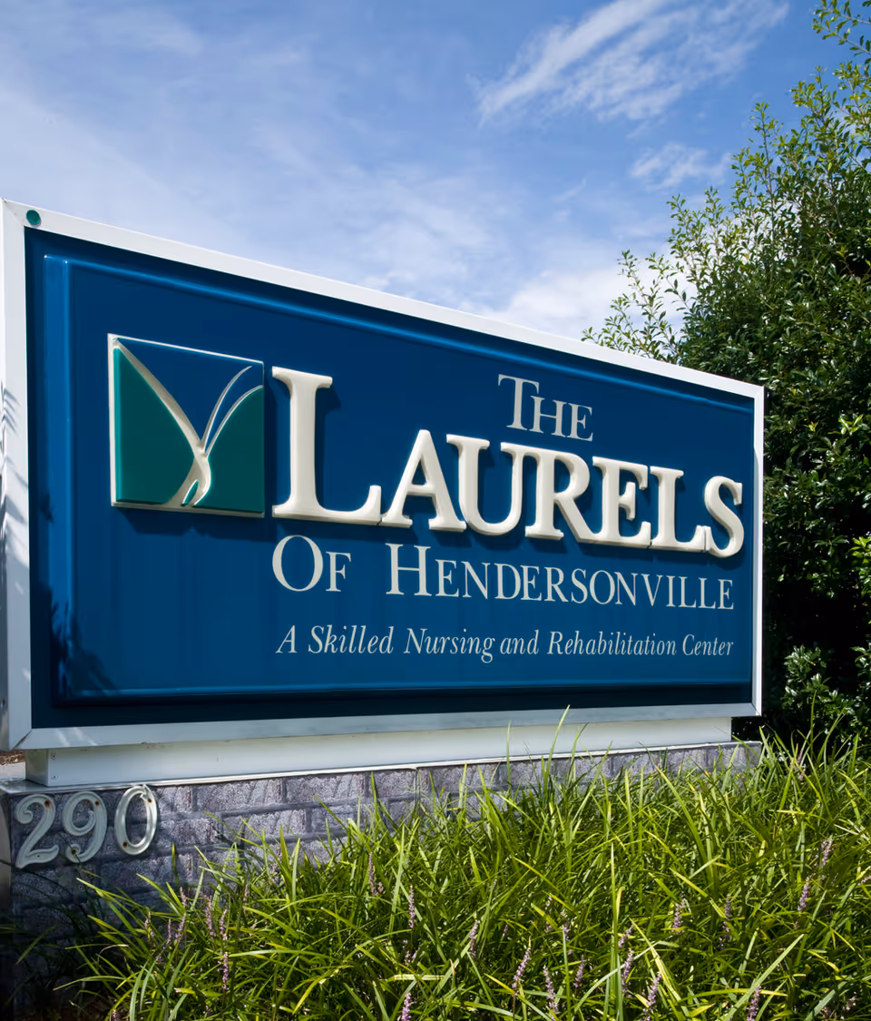 Outdoor sign for The Laurels of Hendersonville, a skilled nursing and rehabilitation center, with green plants in front and a blue sky background.