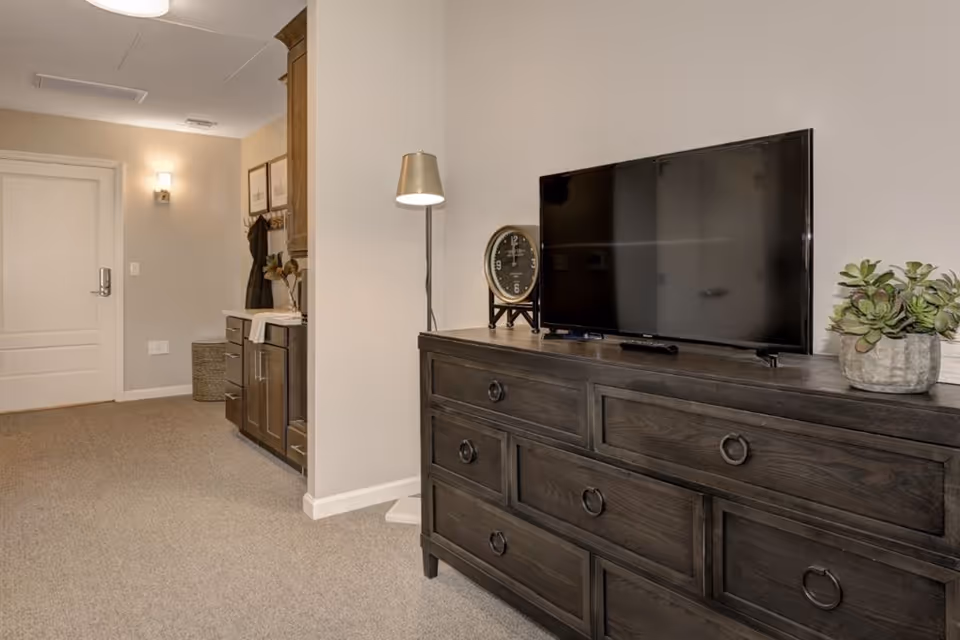 Interior living area with a dark wood dresser topped by a flat-screen TV, lamp, clock and plant, with a view toward an entry door and hallway.