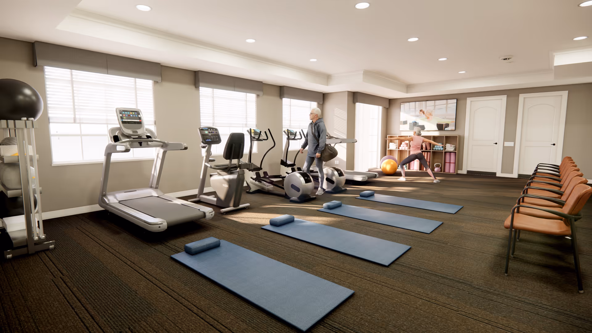 A fitness room with exercise equipment including treadmills, stationary bikes, and elliptical machines. Four blue exercise mats with small blue cushions are laid out on the floor. A man is walking near the exercise bikes, and a woman is stretching near a shelf with exercise balls and towels. There are several brown chairs lined up against the wall on the right side of the room. Large windows allow natural light to fill the space.