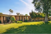Single-story building with a covered patio area and large windows, surrounded by a well-maintained green lawn and various trees under a clear blue sky.