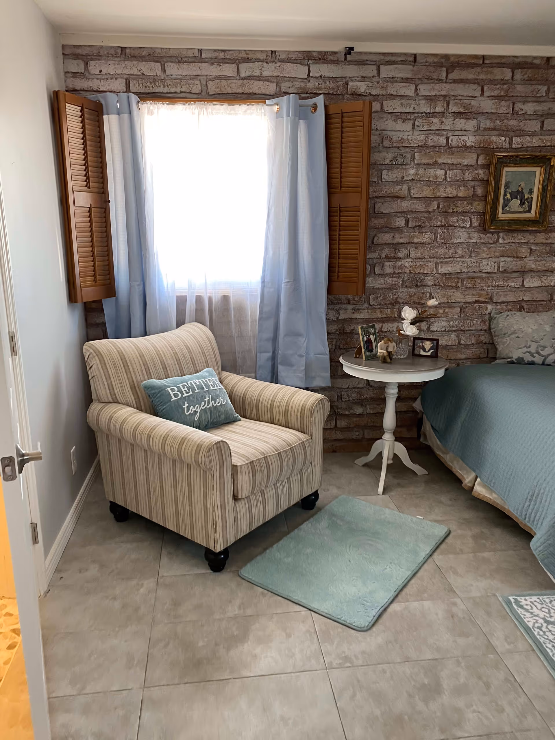 A cozy bedroom corner with a striped armchair featuring a pillow that says 'BETTER together', a small white round side table with framed photos and decorative items, a bed with a teal quilt, and a window with light blue curtains and wooden shutters against a brick accent wall.