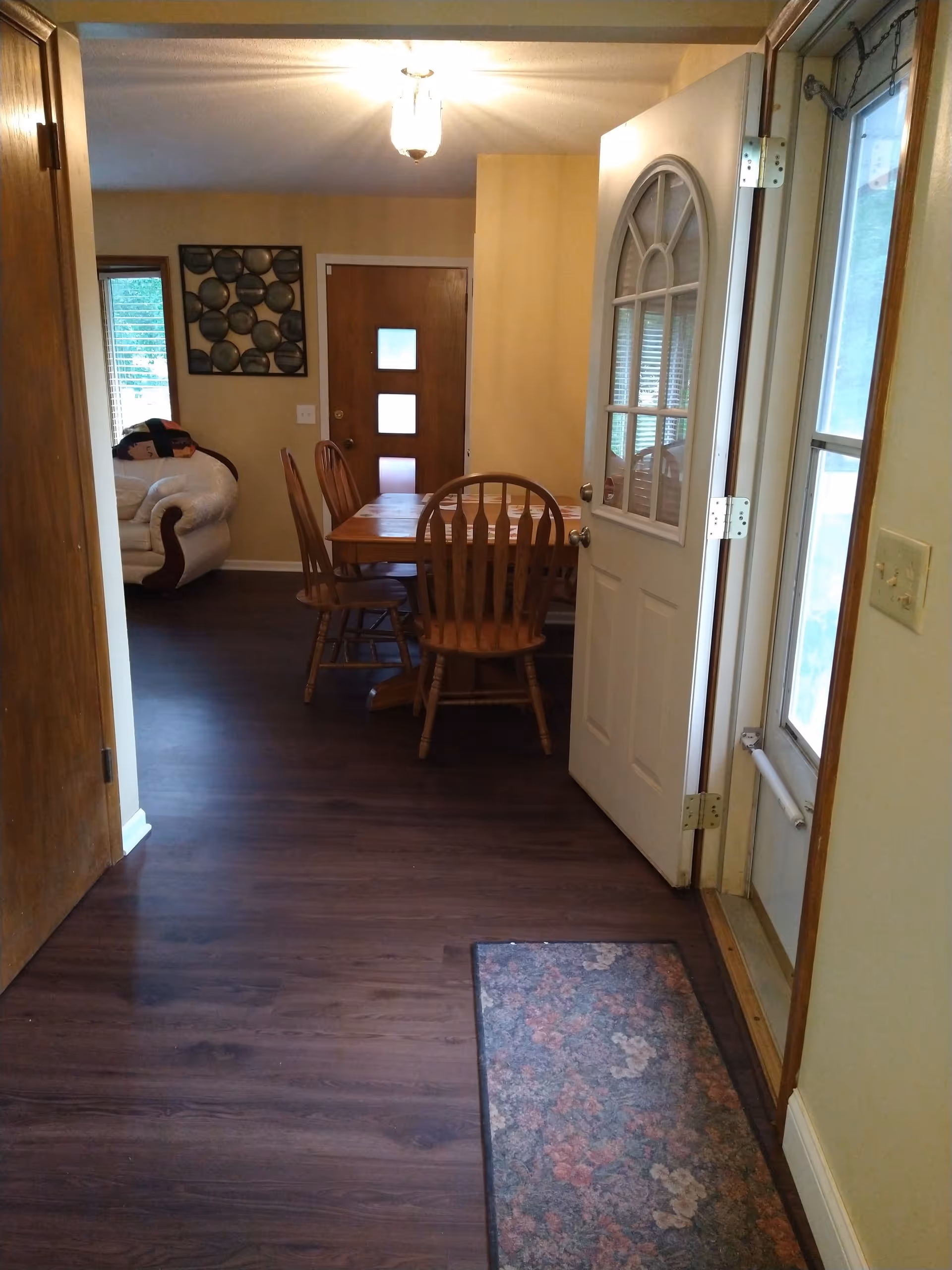 View of an interior space showing a wooden floor, an open white door with a window, a floral rug near the entrance, a wooden dining table with four chairs, a beige wall with a decorative wall art, a window with blinds, and a white cushioned armchair partially visible in the background.