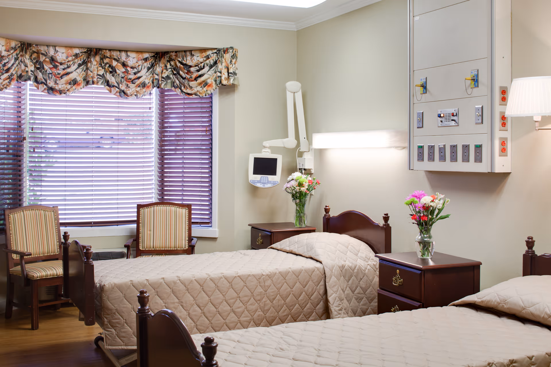 A senior living bedroom with two twin beds, wooden bedside tables topped with vases of flowers, chairs by a window, and medical equipment on the wall.