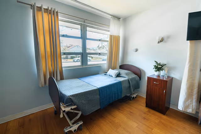 Private room with a single adjustable bed next to a window, a wooden bedside cabinet, and light blue walls.