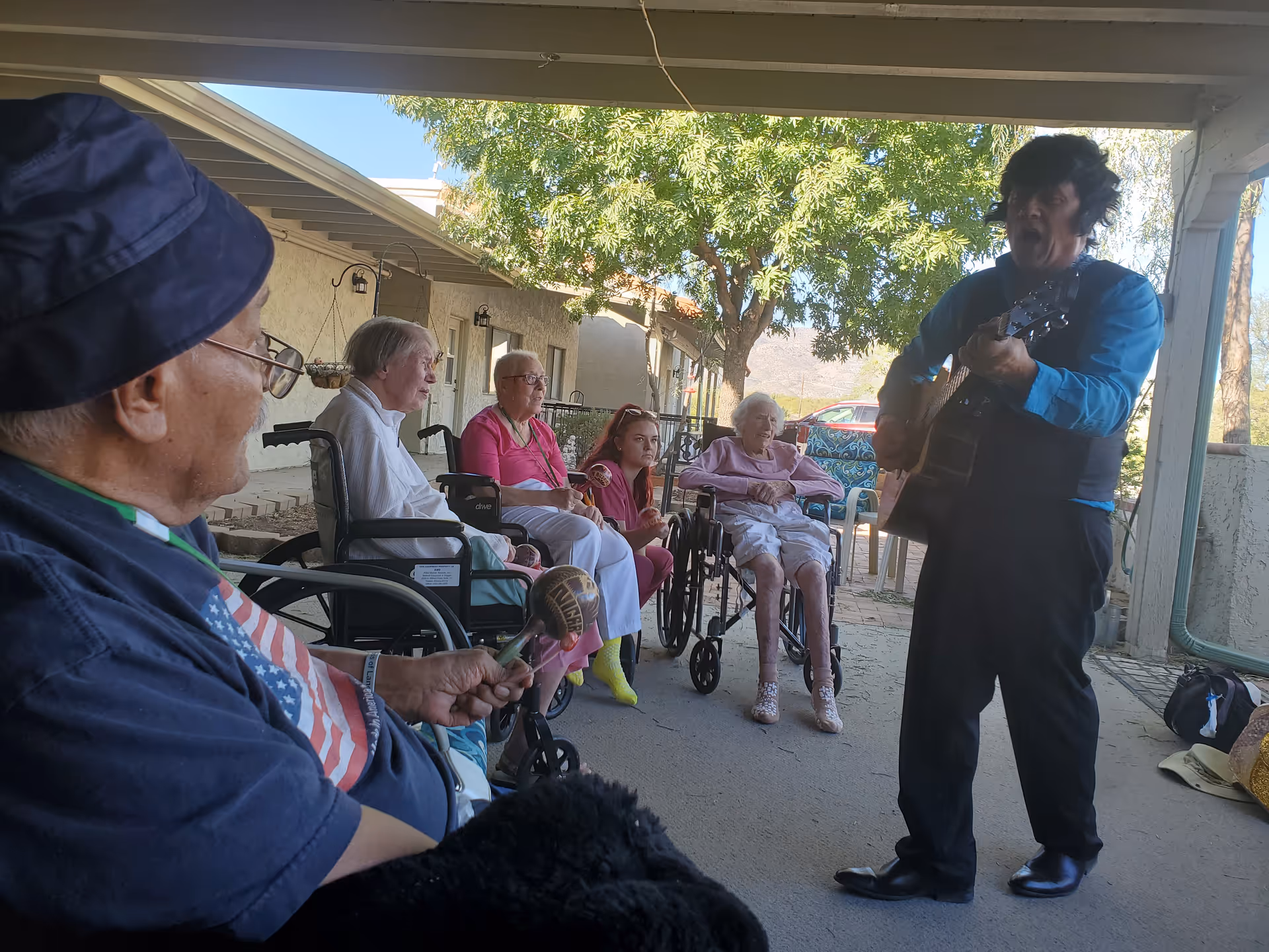 A group of elderly people sitting outdoors under a covered patio, some in wheelchairs, attentively watching a man playing a guitar and singing. The setting is shaded with trees and buildings in the background.