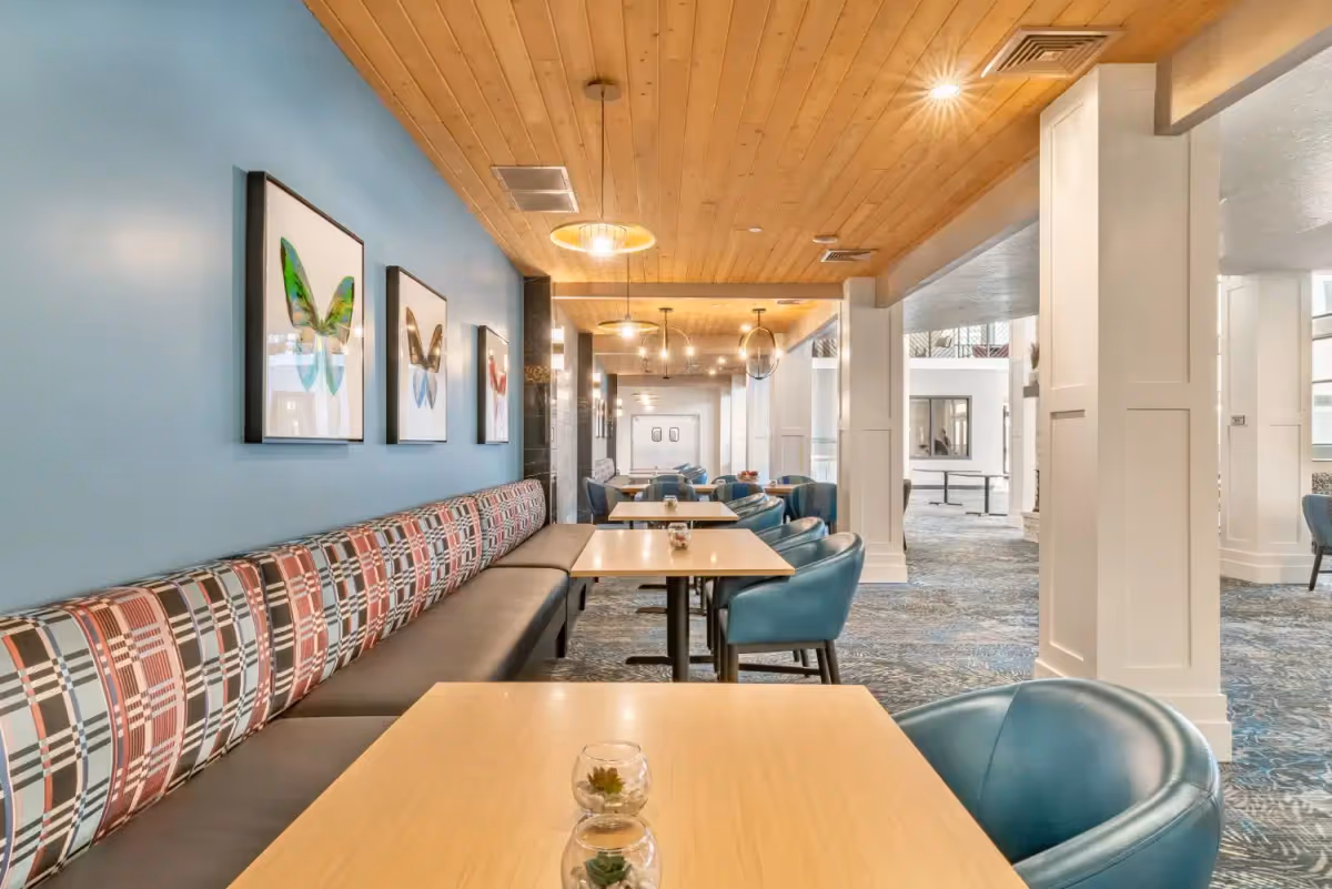 Communal dining area with banquette seating along a blue wall, tables and teal chairs under a wood-paneled ceiling.