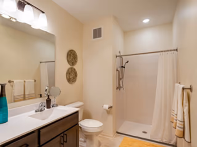A clean bathroom with a white countertop sink, dark wood cabinets, a toilet, and a walk-in shower with a white curtain. There are two decorative round wall hangings above the toilet, a towel rack with towels, and a yellow bath mat on the floor. The lighting is bright with a fixture above the mirror.