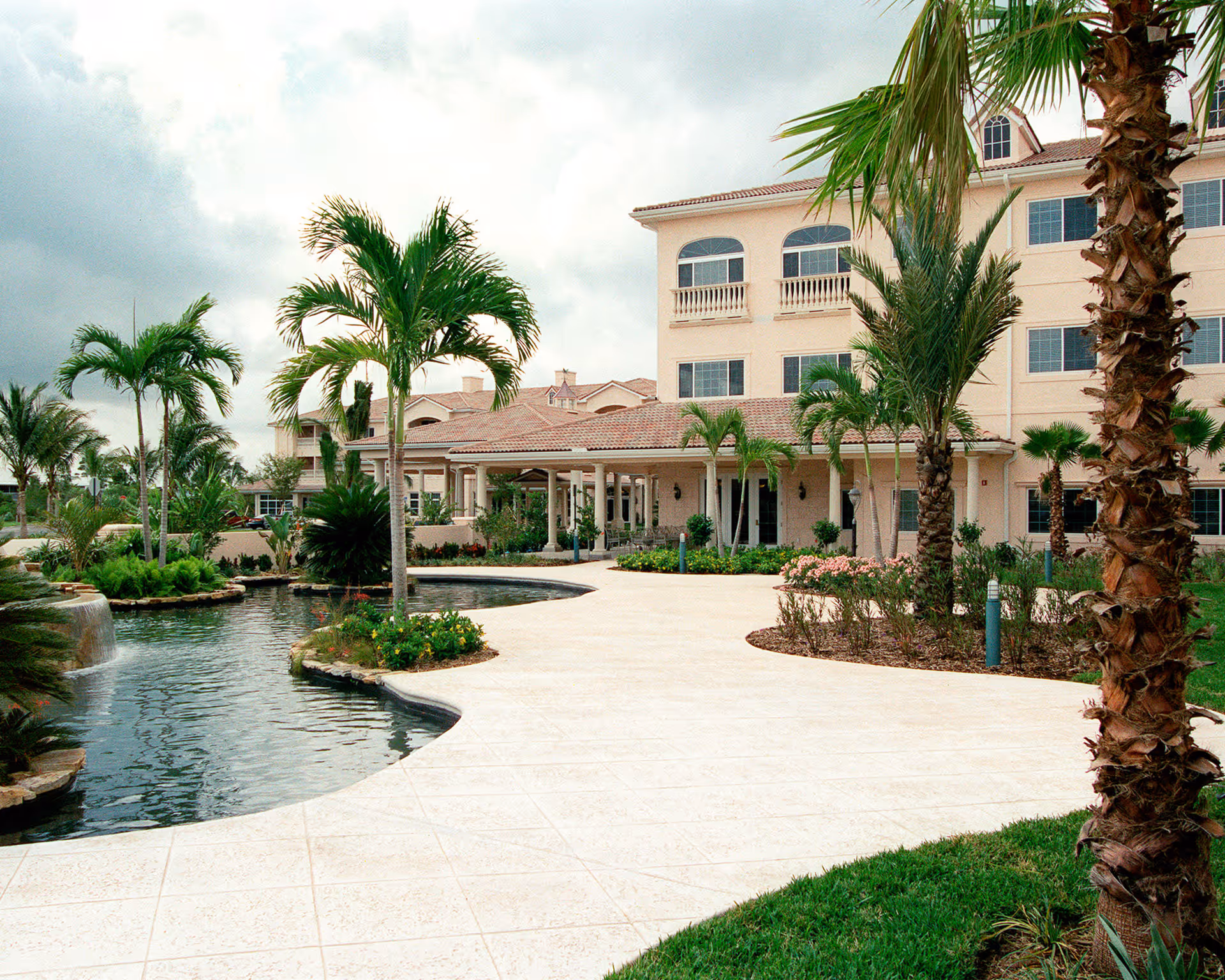 Exterior view of Harbor Place at Port St. Lucie showing a large building with multiple windows, surrounded by palm trees and landscaped gardens. A paved walkway curves around a water feature with a small waterfall.