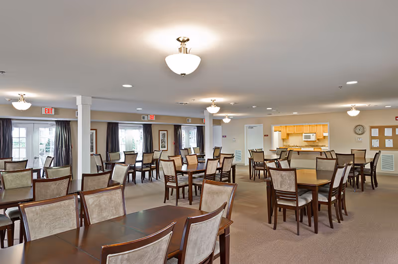 A spacious dining room in a retirement community with multiple wooden tables and cushioned chairs arranged neatly. The room has beige carpeting, white walls, ceiling lights, and large windows with dark curtains allowing natural light. There is a small kitchen area with wooden cabinets and a microwave in the background.