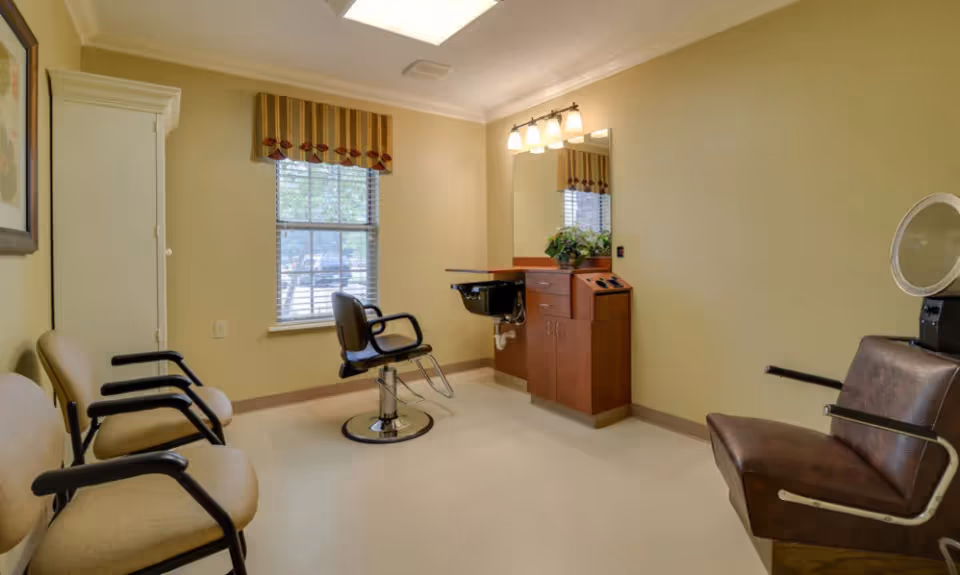 Interior view of a senior living facility's hair salon with a styling chair in front of a mirror and sink, additional seating along the wall, a window with a striped valance, and soft yellow walls.