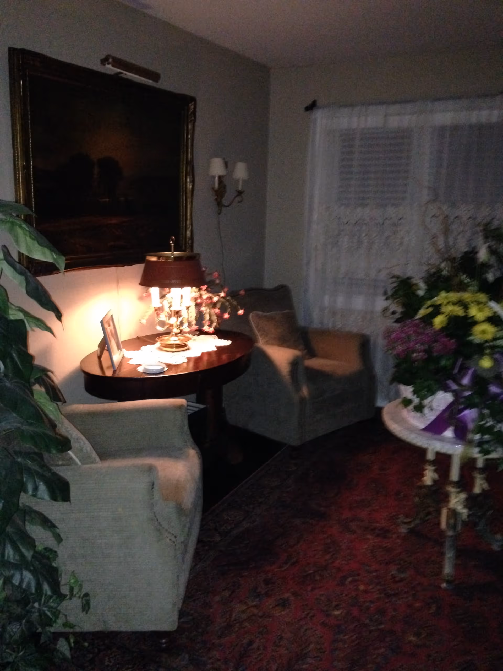 A dimly lit living room corner with two upholstered armchairs, a round wooden table with a decorative lamp and framed photo, a large painting on the wall, a window with sheer curtains, a potted plant, and a small round table with a large floral arrangement on a patterned rug.