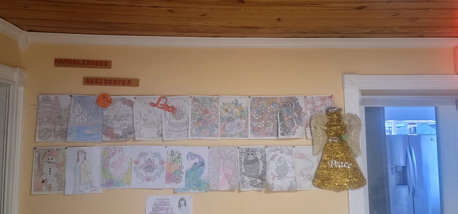 A wall inside a facility with various colored drawings and artwork pinned in two rows. Above the artwork, there are two wooden plaques with the words 'MANUALIDADES' and 'RESIDENTES'. To the right, a decorative golden angel with the word 'Peace' hangs on the wall near a doorway leading to a kitchen area with a refrigerator visible.