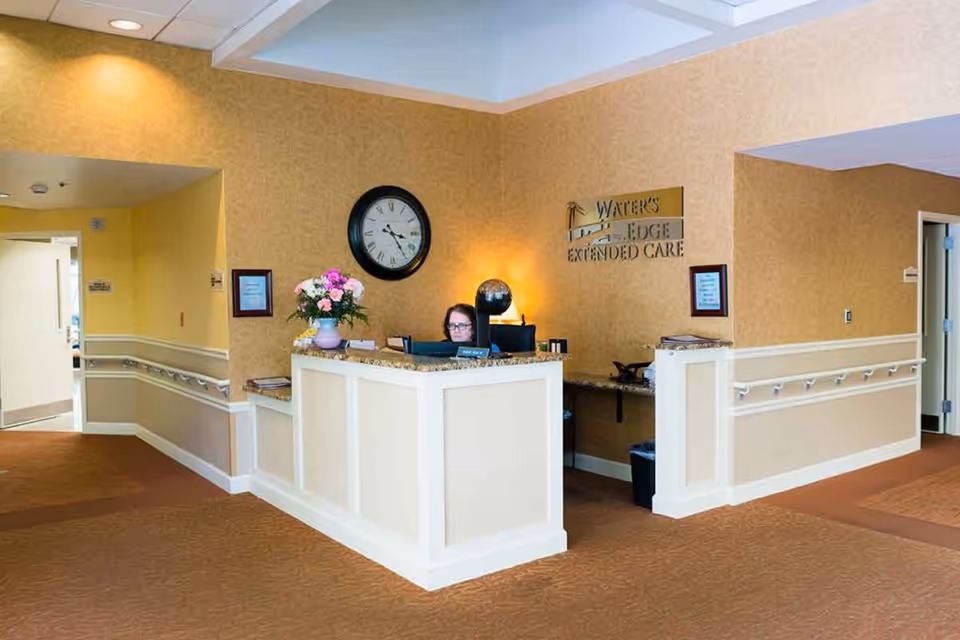 Reception desk area inside Water's Edge Extended Care facility with a staff member seated behind the desk. The walls are painted yellow with white trim, and there is a large clock on the wall above the desk. A flower arrangement is placed on the desk, and the facility's name is displayed on the wall behind the desk.