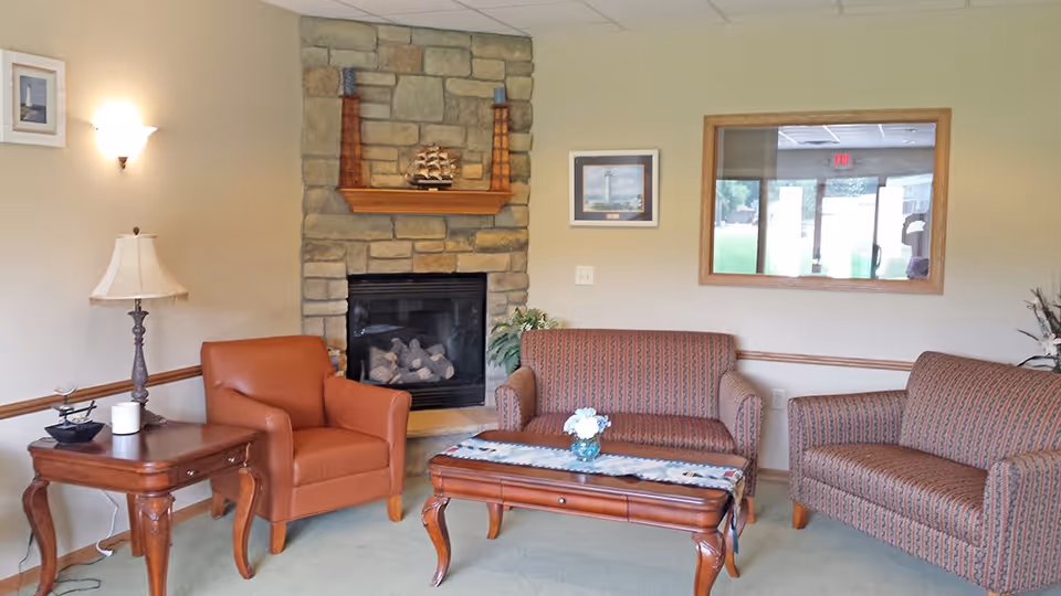 A cozy living room area with a stone fireplace in the center. There is a brown leather armchair to the left, two patterned fabric sofas on the right, and a wooden coffee table with a small flower arrangement in the middle. A wooden side table with a lamp and decorative items is next to the armchair. The walls are beige with framed pictures and a window showing an outside view.