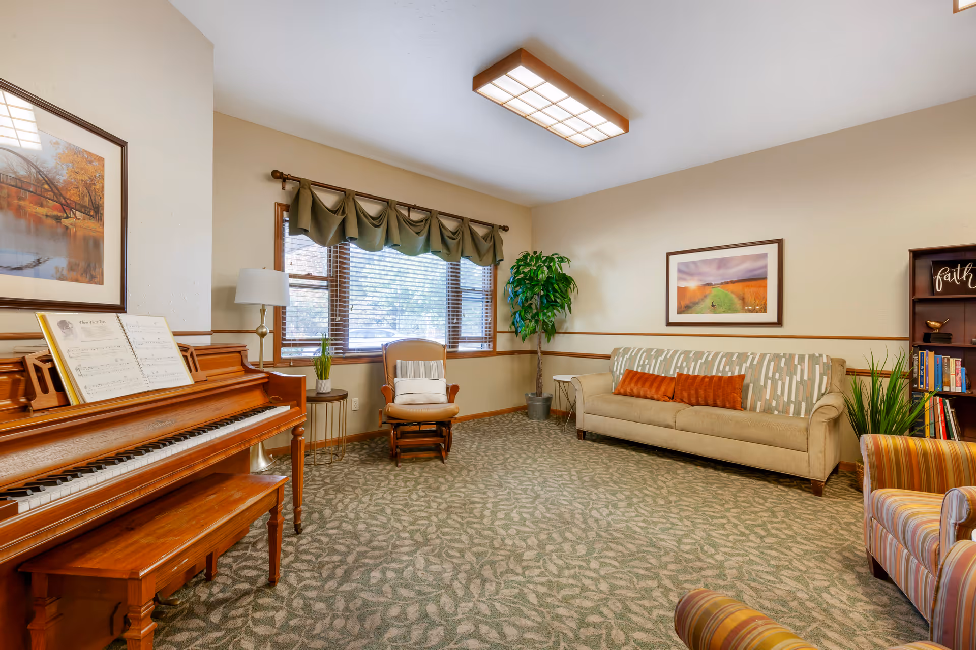 A cozy common living room with a piano, upholstered sofa and armchairs, a rocking chair by a window, potted plants, and framed artwork on the walls.