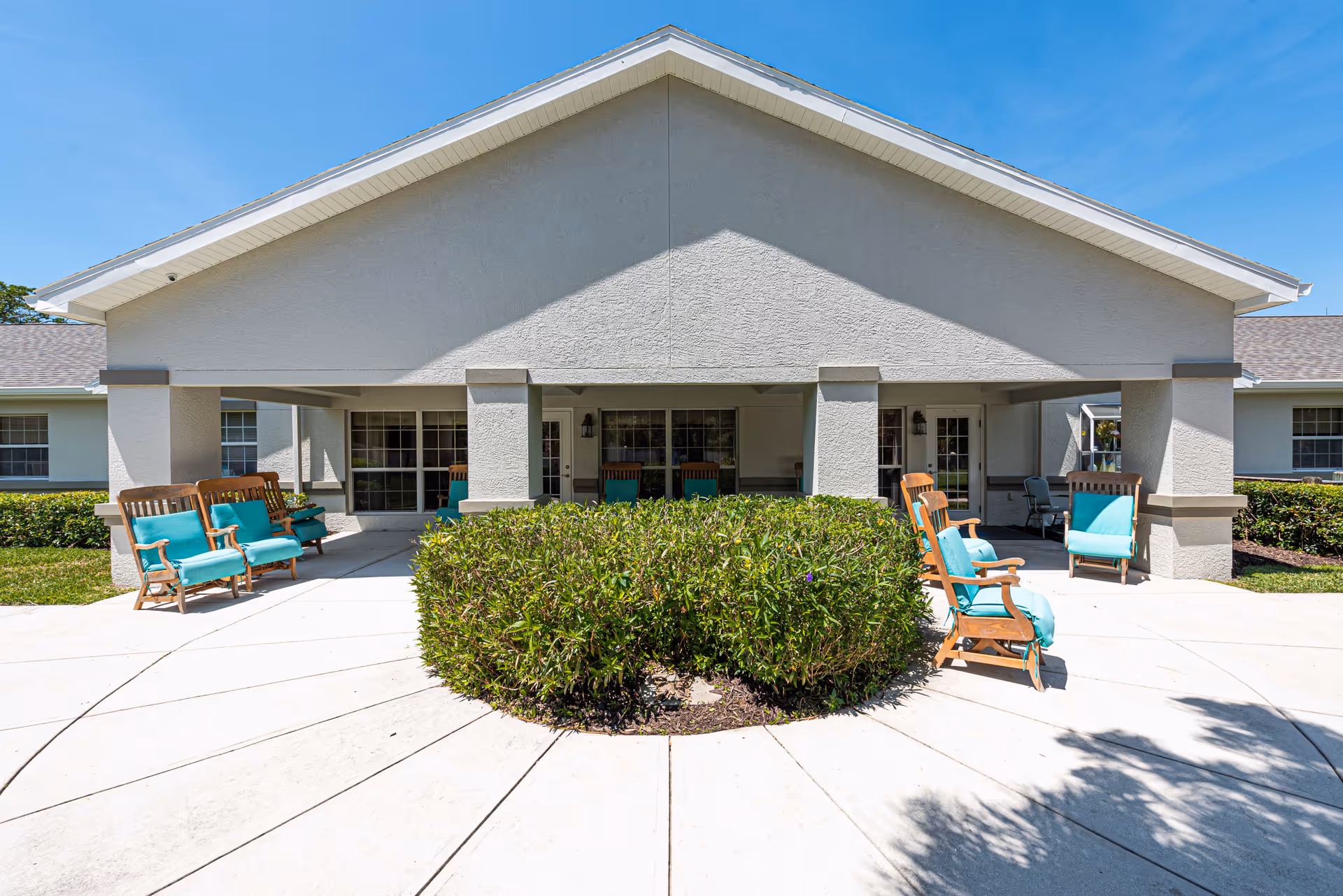 Exterior view of a single-story building with a covered patio area featuring wooden chairs with teal cushions. There is a circular hedge in the center of a concrete patio under a clear blue sky.