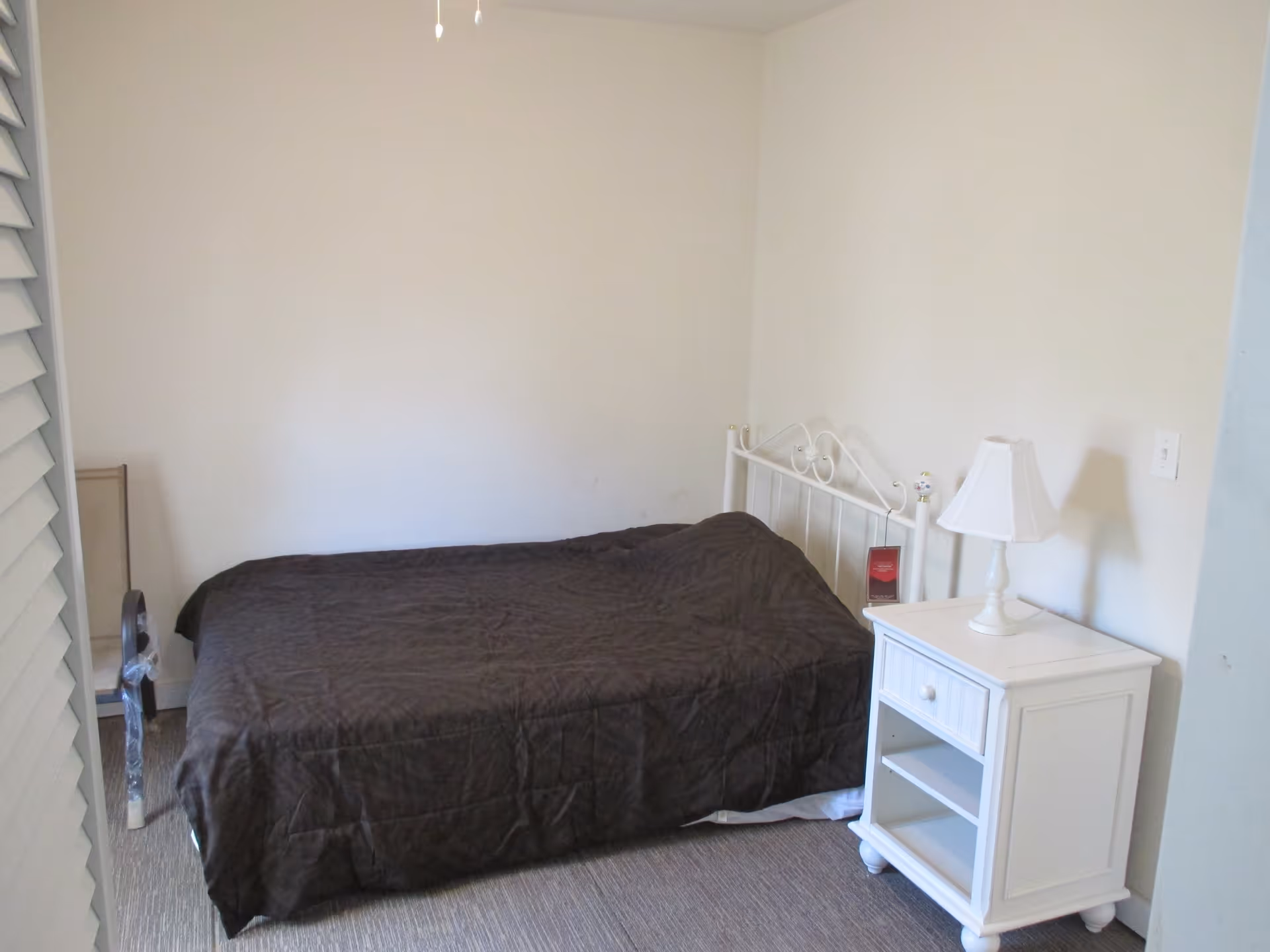 A simple bedroom with a single bed covered in a dark brown quilt, a white metal headboard, and a white nightstand with a white table lamp on it. The walls are plain and light-colored, and there is a carpeted floor.
