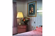 Cozy bedroom corner with a bed, a nightstand holding a lamp and a vase of flowers, and framed artwork on patterned wallpaper.
