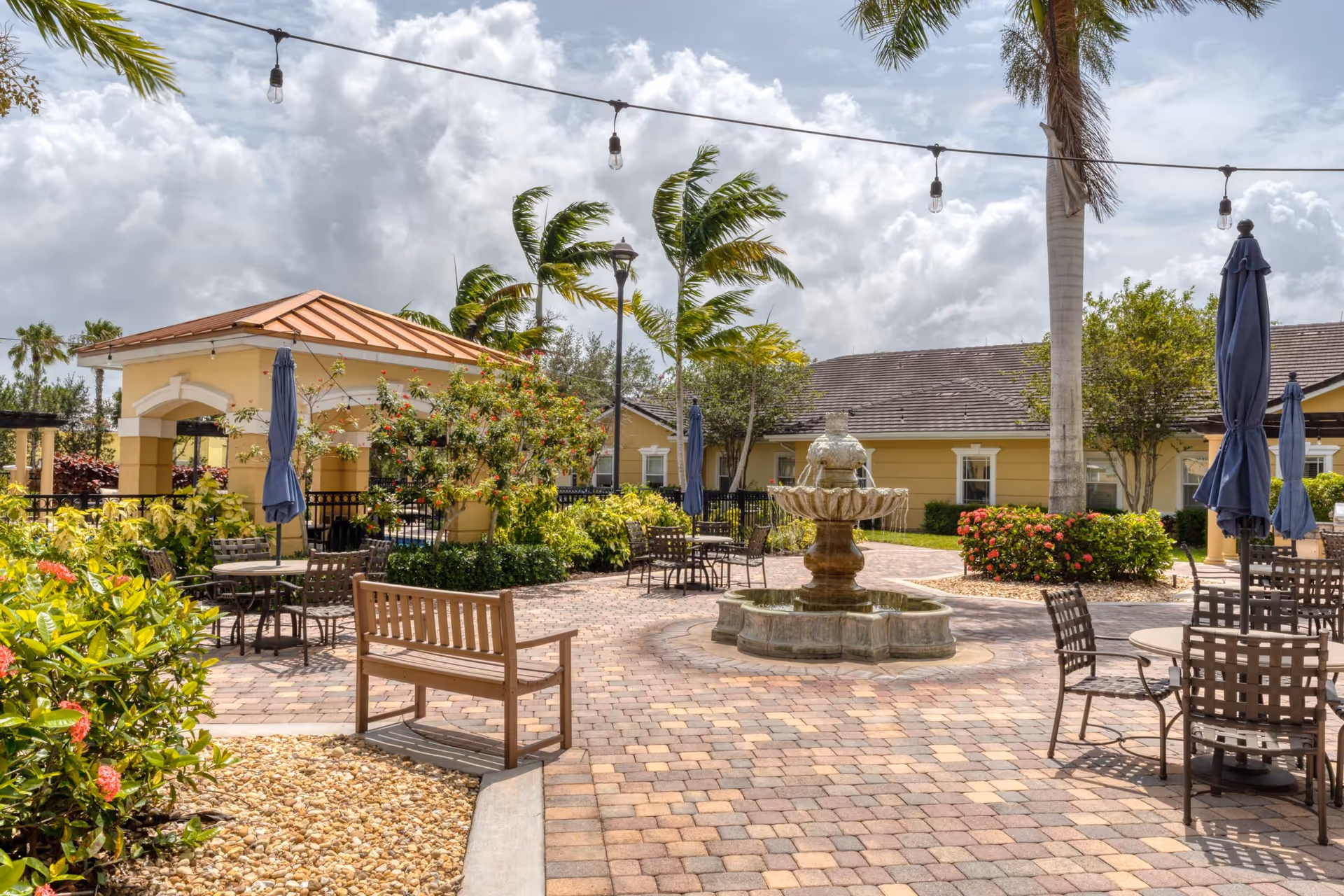 Outdoor courtyard area with a stone fountain in the center, surrounded by patio tables with umbrellas, a wooden bench, lush greenery, and palm trees under a cloudy sky.