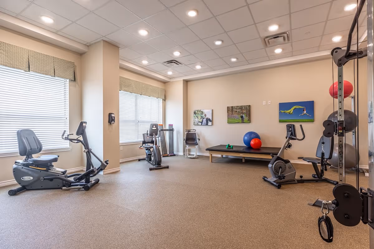 A well-lit exercise room with two recumbent stationary bikes, a weight machine, a bench with exercise balls and dumbbells, and three framed pictures on the wall. The room has large windows with blinds and beige walls.