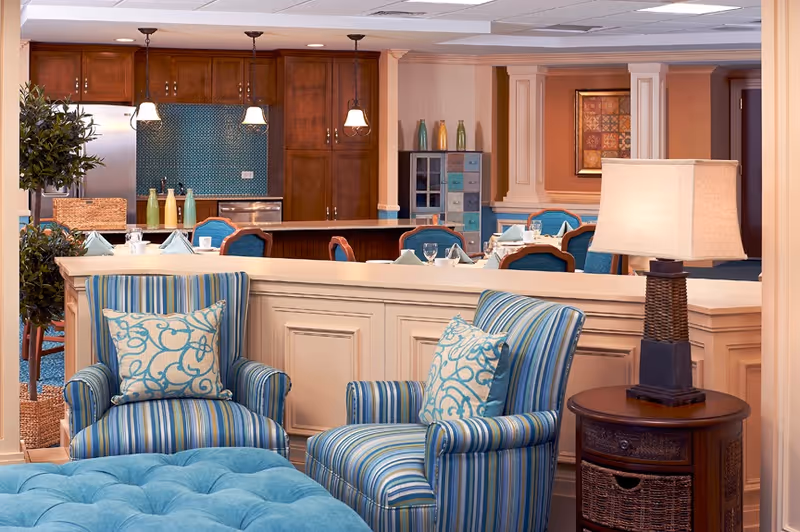 A cozy interior space featuring two striped armchairs with patterned cushions, a blue tufted ottoman, and a wooden side table with a lamp. In the background, there is a dining area with tables set with napkins and glasses, and a kitchen area with wooden cabinets and a blue tiled backsplash.