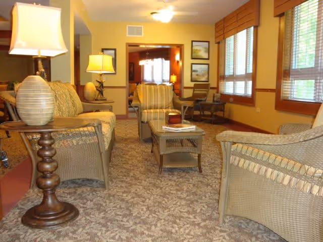 A cozy living room area with wicker furniture including armchairs and a sofa, a wooden coffee table with books on it, two table lamps with beige lampshades, carpeted floor with a patterned design, yellow walls, and large windows with wooden blinds letting in natural light.