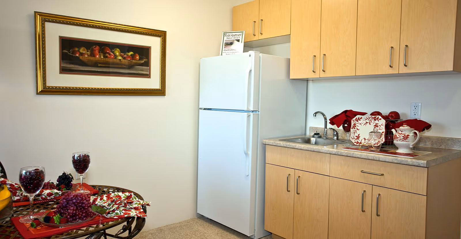 A small kitchen area with light wood cabinets, a white refrigerator, a countertop with a sink, and red and white decorative dishware. A framed picture of a bowl of apples hangs on the wall. A round table with a glass top holds two glasses filled with grapes and a plate of grapes with a red napkin.