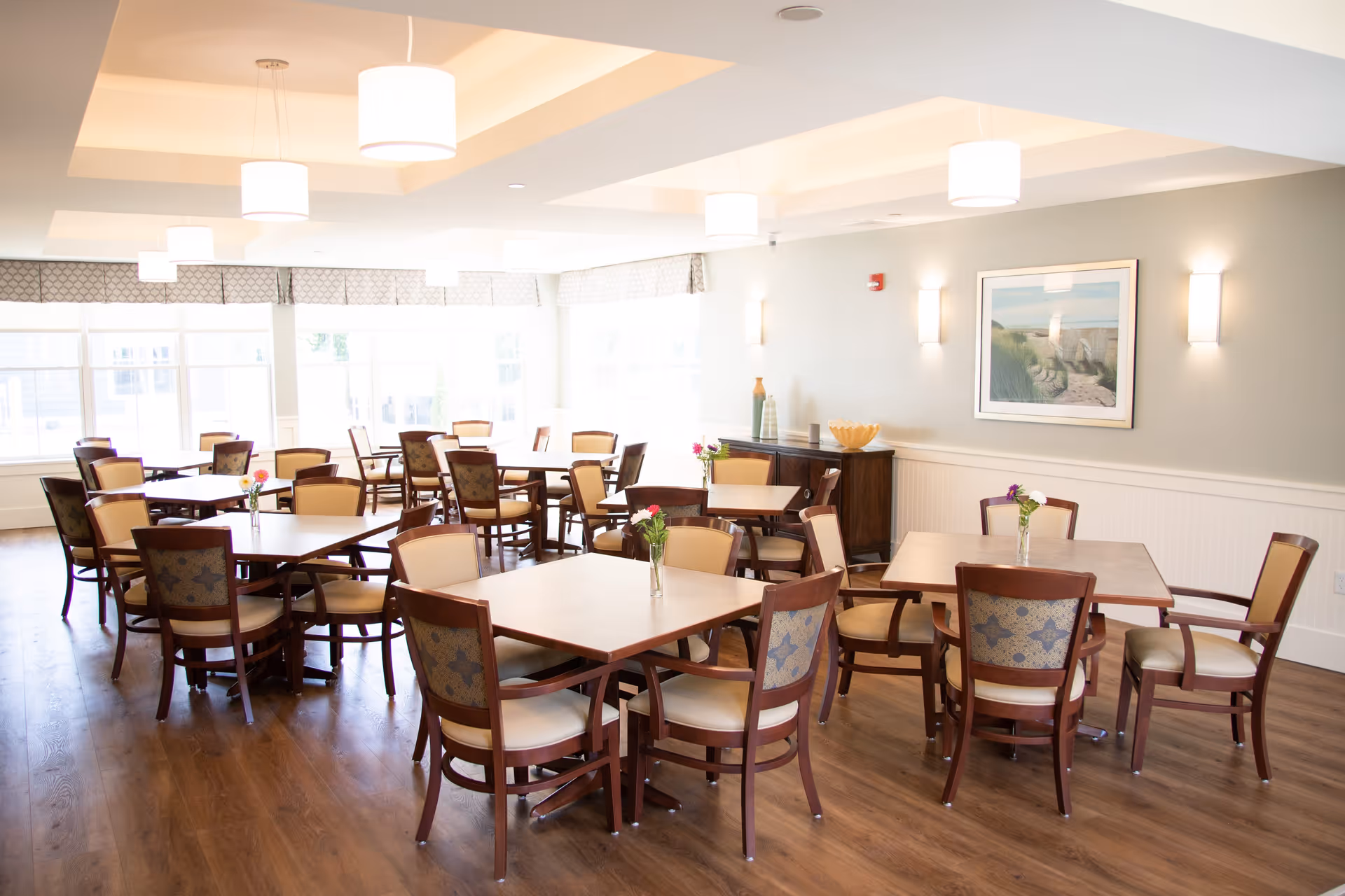 A bright dining room with multiple wooden tables and chairs arranged neatly. Each table has a small vase with flowers. The room has large windows letting in natural light, light-colored walls, and ceiling lights. A framed picture hangs on the wall above a sideboard with decorative items.