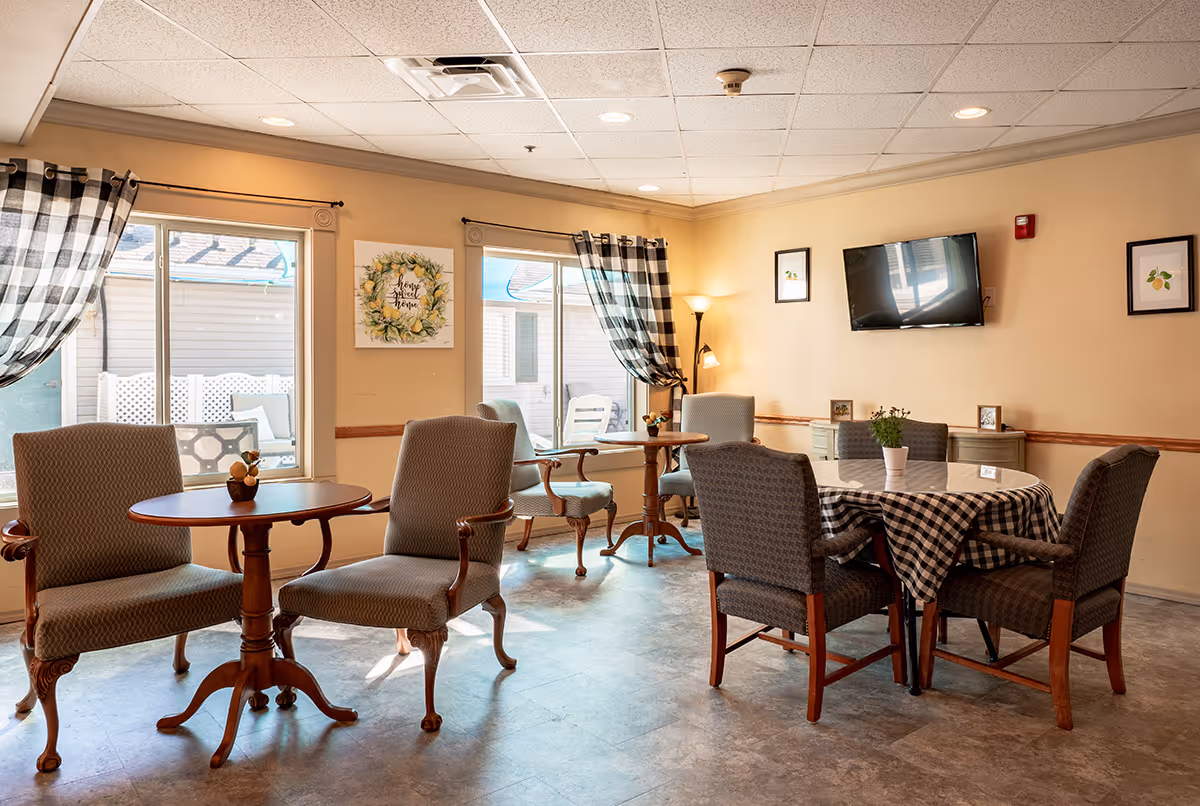 A cozy common area with several small tables and cushioned chairs arranged around them. The room has large windows with checkered curtains allowing natural light to enter. A round table with a checkered tablecloth and potted plant is in the foreground. The walls are decorated with framed pictures and a wall-mounted TV. A floor lamp provides additional lighting.