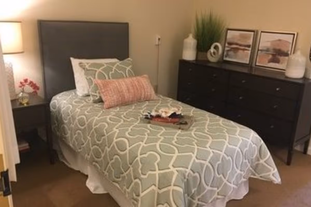 A neatly made single bed with a gray patterned bedspread and multiple pillows, including a pink accent pillow. Next to the bed is a dark wooden nightstand with a lamp and a small vase with pink flowers. On the right side of the room is a dark wooden dresser with decorative items including two framed pictures, a plant, and white vases.