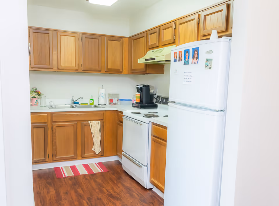 A small kitchen with wooden cabinets, a white refrigerator with photos and notes attached, a white stove with an overhead vent, a coffee maker, a sink with a dish soap bottle, and a striped red and beige rug on a wooden floor.