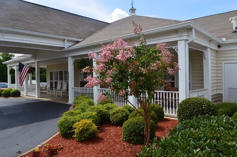 Exterior view of a single-story building with a covered porch featuring white railings and rocking chairs. There is a well-maintained garden with green shrubs and a flowering tree with pink blossoms in front of the porch. An American flag is displayed near the entrance.