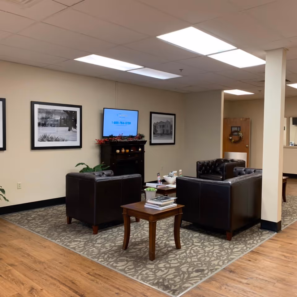 A lounge area with dark leather armchairs around a coffee table, a wall-mounted TV, framed photos, and a patterned rug.